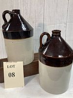 Two antique whiskey jugs, brown top and tan base sitting side by side with lot number card