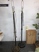 Photo showing two floor lamps, one black and one brass tone, with metal bases featuring grid and pattern details, both lamps with electrical cords and exposed bulbs, no lampshades.