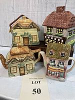 Front view of four ceramic items shaped as cottages and shops with distinct roofs and detailing. Shows two teapots and two biscuit jars in earthy painted tones.