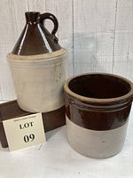 Full view of one gallon brown and tan whiskey jug with handle beside two-tone brown and tan one gallon crock on wooden block against wooden backdrop.