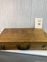 Rectangular wooden briefcase box closed, showing smooth top surface and handle with metal latches on front panel.