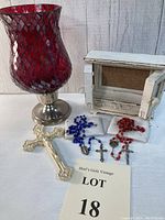 Photo showing red glass candle holder in silver base, white wooden picture frame with cork board, two glass rosaries in blue and red with white zipper pouches, and small white ceramic wall cross.