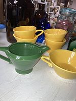 Yellow ceramic cups stacked, green ceramic creamer with spout in front, arranged with other glass bottles in background.
