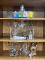 Full view of vintage glassware collection on shelf including branded cans, various decanters, pitchers, and glasses arranged on two wooden shelves