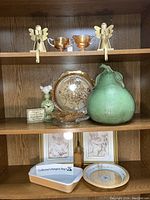 Side view of wooden shelf containing carnival amber glassware and green decor items including pear sculpture, clock, and framed cherub prints