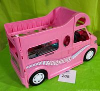 Side and angled views of a pink plastic toy camper van with 'ADVENTURE' printed on the side, showing front and rear with interior details such as a stove and steering wheel with sound buttons.