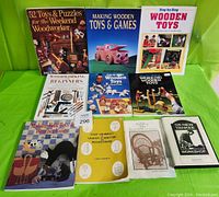 Photo showing 9 books arranged in two rows on a green surface, titles visible include 52 Toys & Puzzles for the Weekend Woodworker, Making Wooden Toys and Games, Step-By-Step Wooden Toys, Woodworking for Beginners, Wooden Toys Puzzles and Games, Shop Drawings of Shaker Furniture and Woodenware, Building the Bent Willow Chair, The New Yankee Workshop CD case.