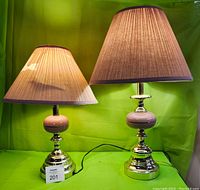 Two brass table lamps side by side on a green background, both with pink ball accent and fabric lampshades; one shade appears uneven, consistent with damage.