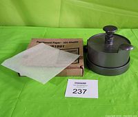 Metal hamburger press with box of parchment paper sheets and one sheet displayed on top of the box.