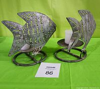 Two metal fish-shaped candle holders on a green background, each with one white votive candle placed on round bases.