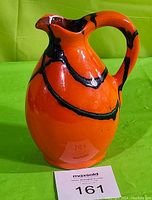 Front and side views of an orange and black ceramic pitcher jug on a green background, showing shape, color, and pattern design.