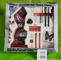 Front of boxed microscope set showing red microscope, projector attachment, several accessories and specimens included, within original foam packing.