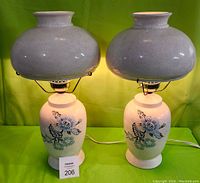 Pair of white ceramic table lamps standing side by side, showing overall design and shape of base and shade against green background.