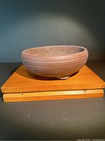 Front view of a round bonsai pot on a wooden stand showing its purple terra cotta clay and three feet.
