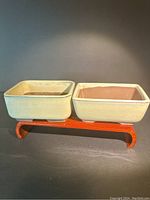 Two rectangular light glazed bonsai planters on a small wooden display stand, showing front and side views, highlighting their shape, glaze, and footed bases.