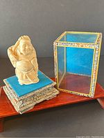 Full view of Buddha figurine on blue fabric base, next to the clear glass display box with floral trim