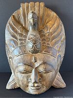 Front view of large hand carved wooden Buddha mask with peacock motif on headdress.