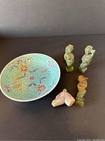 Photo showing three jade figurines, one famille rose porcelain plate, and a stone netsuke dog's head arranged on a dark surface.