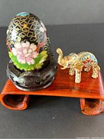 Photo showing the black cloisonné egg with pink flower designs and the miniature cloisonné elephant figurine on small wooden stands.