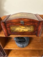 Front view of the curved-top wooden box with brass panel and metal clasp on a wooden shelf.