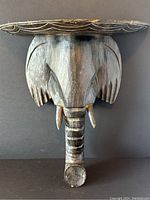 Front view of the carved wooden elephant head with a decorative shelf above it, showing the intact tusks and striped trunk.