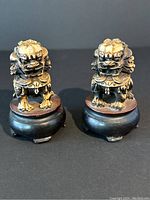 Front view of pair of small resin foo dog statues on round wooden stands showing detailed front faces and paws.