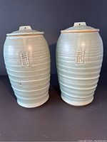 Front view of two greenish blue soft glazed ceramic pots with lids on, both showing raised rectangular markings and ribbed horizontal texture.