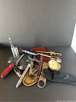 Overview of men's lot showing leather wallet, passport folio, pens, multi-tools, watch, travel alarm clock, brass tray, and cigar tube