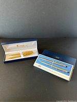 Two boxed pen and pencil sets: one Bill Blass gold-tone set and one silver-tone Cross set side by side.