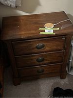 Side view of solid wood side stand with three drawers and metal ring pulls on a carpeted floor, with some wear marks visible on the top surface.