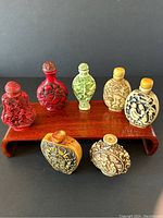 Full view of set showing seven Chinese snuff bottles of various carved materials and colors on a wooden stand. Two red cinnabar bottles, two missing tops, two adult-themed bottles visible.