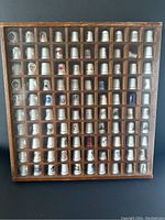 Full display case showing 100 porcelain thimbles arranged in a wooden compartmentalized case with a transparent front.