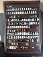 Full view of wood and glass display case filled with rows of 163 assorted decorative thimbles of mixed materials and styles.