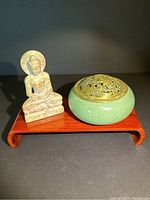 Photo of carved stone Buddha statue and green crackle glazed incense burner set on a small wooden stand against a dark background