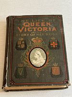 Front cover of 1901 book "The Life of Queen Victoria and the Story of Her Reign" showing decorated embossed cover with orange title, various coat of arms crests, and oval portrait of Queen Victoria.