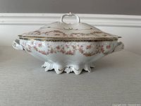 Porcelain tureen with floral bridal rose design, gold trim, and ornate base and handles shown from front with lid on.