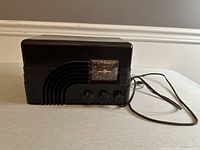 Front view showing black tabletop radio with art deco design and three control knobs with power cord coiled to side