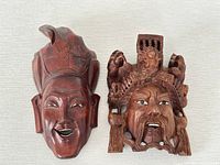 Pair of Chinese wood carved masks, one with a smiling face and headdress and the other with an elaborate headdress and solemn expression shown together on a light background.