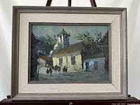 Framed oil painting showing a historic village scene with church and horses, displayed on an easel.