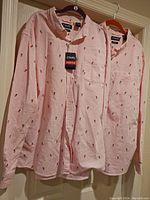 Two men's pink and white striped button-down shirts with small pineapple print, hanging on black and orange hangers. One shirt has tags attached.