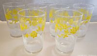 Five clear vintage juice glasses displayed showing yellow floral bands and frosted geometric patterns. All glasses appear intact with no chips or cracks.
