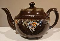 Brown ceramic teapot with a glossy finish, hand painted detailed floral design in multiple colors and gold accents, close-up of body showing decorations.