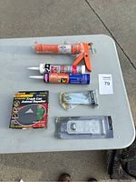 Photo displaying all items on a grey table including caulk gun, three tubes of caulk/sealant, trash can animal repeller, brass faucet valve, and stainless steel latch.