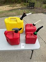 Four plastic fuel cans on a white table outdoors: one yellow diesel can and three red gasoline cans, each with pouring spouts attached.