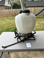 Front view of large white translucent backpack sprayer tank with black lid and metal frame base on grey table.