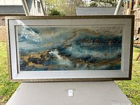 Full view of the large rectangular framed artwork depicting abstract waves in blue, white and gold hues, set in a light wooden frame with white matting.