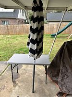 Photo of closed black and white striped patio umbrella standing beside a white folding table outdoors on a concrete surface with a backyard and fence in the background.