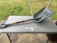 Large scoop shovel resting on a table, showing entire shovel including the plastic scoop and wooden handle with D-grip.