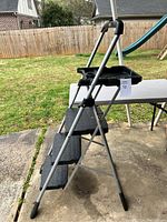 Side view of the large step ladder showing the three wide steps and handle, placed outdoors on concrete with grass and wooden fence in background.