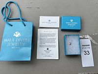 Lot items arranged: Maui Divers Jewelry branded blue bag, informational card, business card, blue box containing pearl pendant on foam insert, and a MaxSold auction tag.
